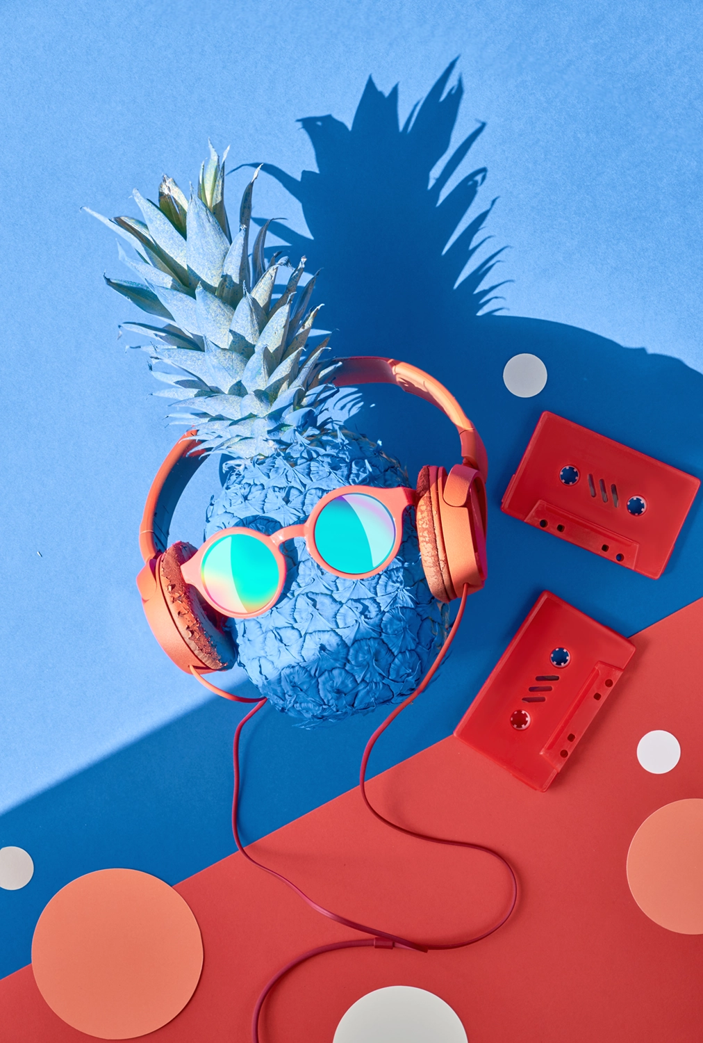 image composée d'un ananas bleu avec des écouteurs rouges et des lunettes fun, des cassettes audio rouges déposées à côtés