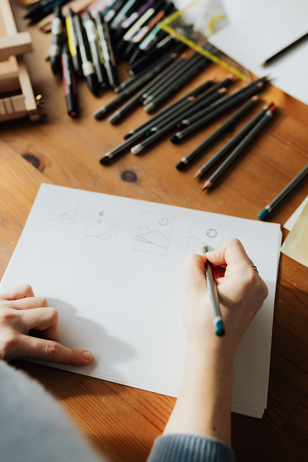 Photo avec vue sur la main droite d'un graphiste qui dessine sur une feuille de papier avec des crayons à papier tout autour de lui.