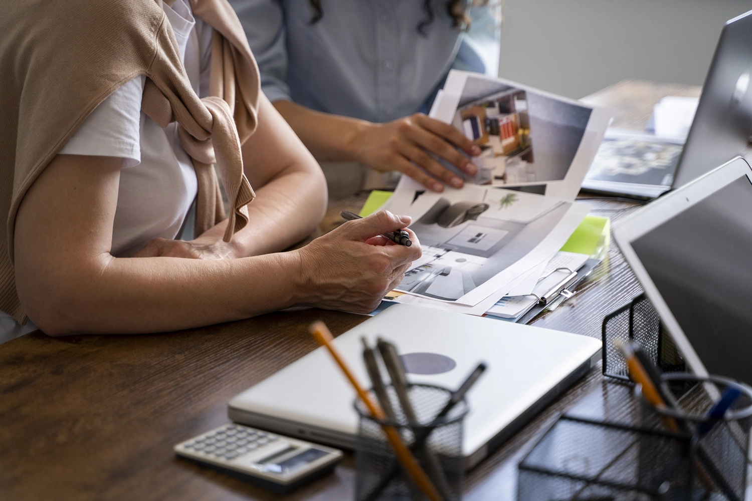 Photo d'une vue au premier plan d'un ordi portable sur un bureau avec un pot de crayon et une femme les bras sur le buereau un stylo en main puis une autre personne à côté d'elle entrain de travailler